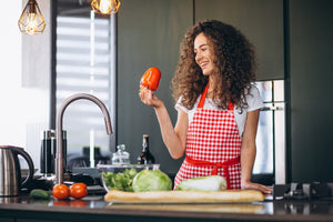 Woman in a kitchen holding a red pepper, surrounded by vegetables.